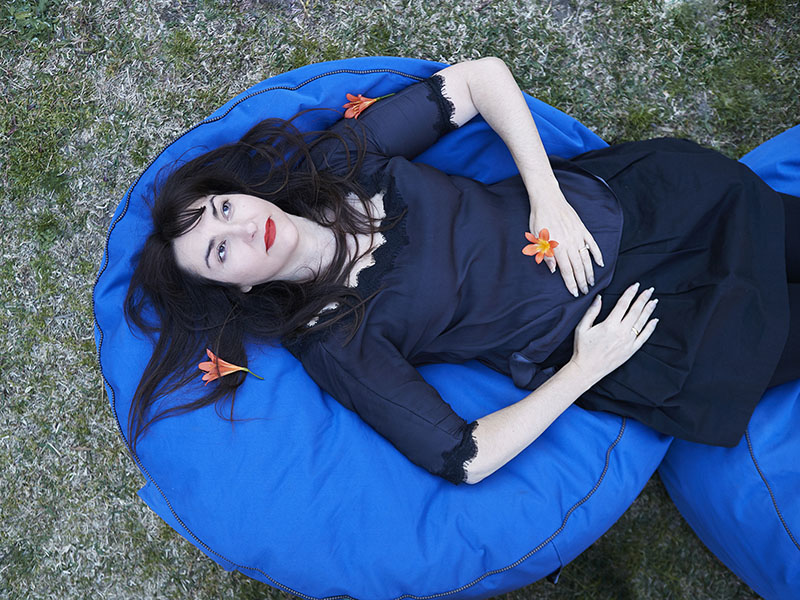 Nike Savvas, dressed in a dark blue dress, lies on a lighter blue beanbag on the grass, holding and surrounded by orange flowers