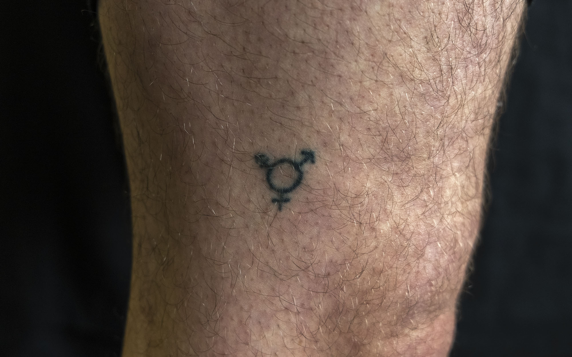 Close up of the side of a leg with a tattoo of a circle and three symbols coming out of it: the female symbol, the male symbol, and a combination of the two