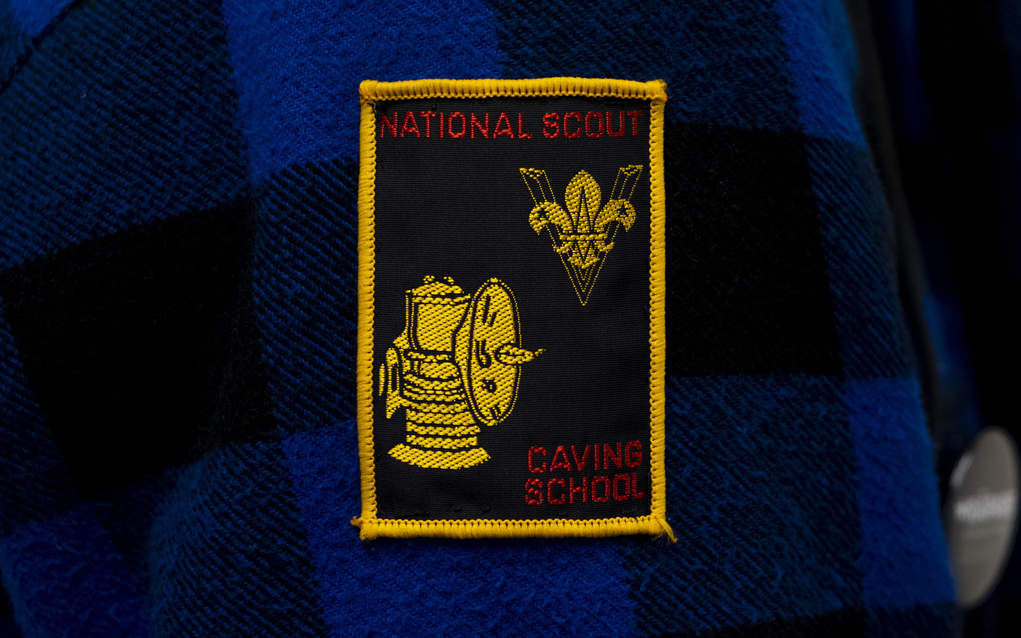 A badge sewn onto a black and blue checked woollen jacket. The badge has a bright yellow border and says "National scout, caving school" on it with a picture of a lantern and the Scouts logo