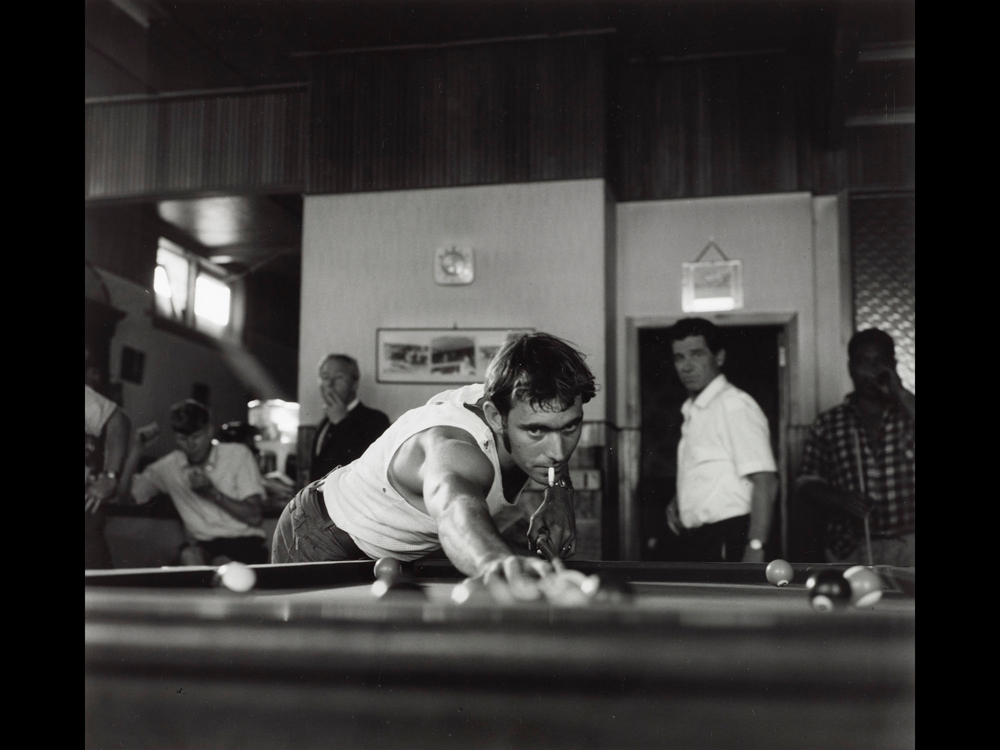 Low-angle shot of a man with a cigarette in his mouth playing pool while other men stand around behind him