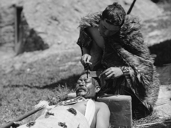 A black and white photograph of a Maori man in a cloak chiseling a tā moko onto the face of another man