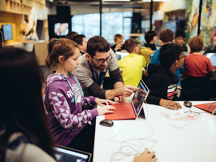 A man shows a young girl something on a computer in busy room