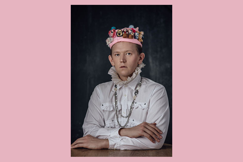 Portrait of Chris Parker wearing a felt hat. The hat features tiny versions of the key New Zealand public figures during Covid-19, a bag of flour, a tiny self-portrait of Chris wearing his orange hoodie, and a tiny official Covid-19 symbol