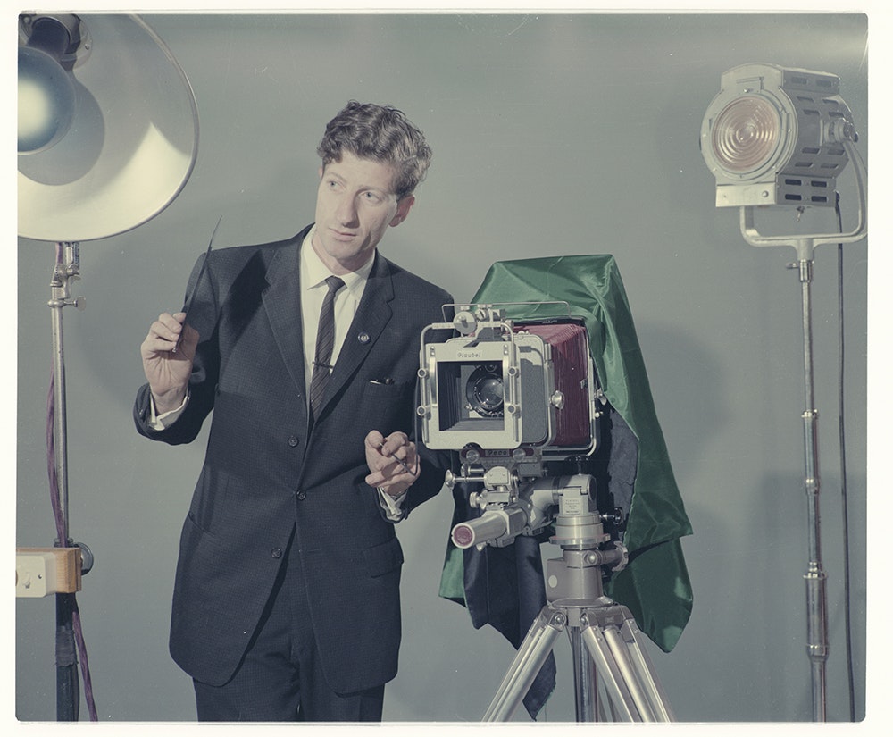A man in a suit stands between stage lights and has an old-fashioned camera on a tripod next to him