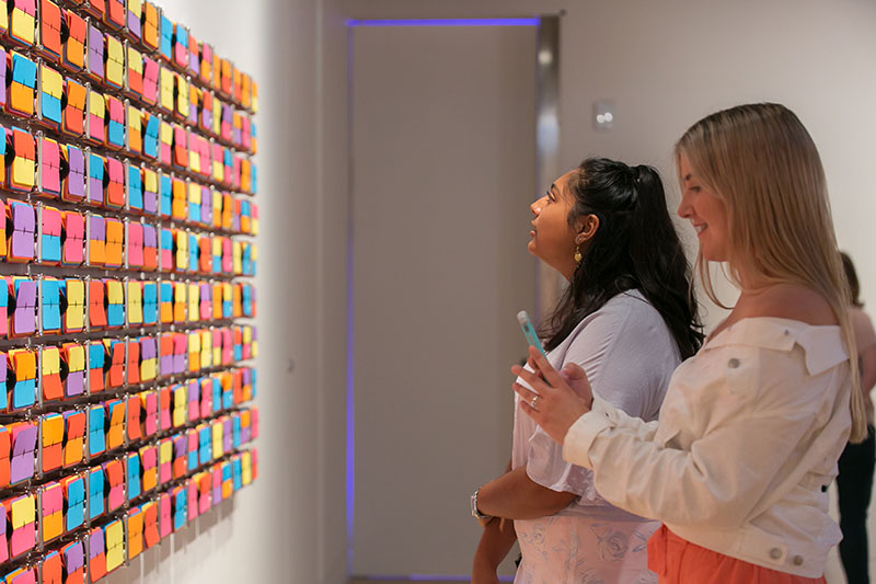 Two women stand in front of Rebecca Baumann’s Automated colour field (Variation 8)