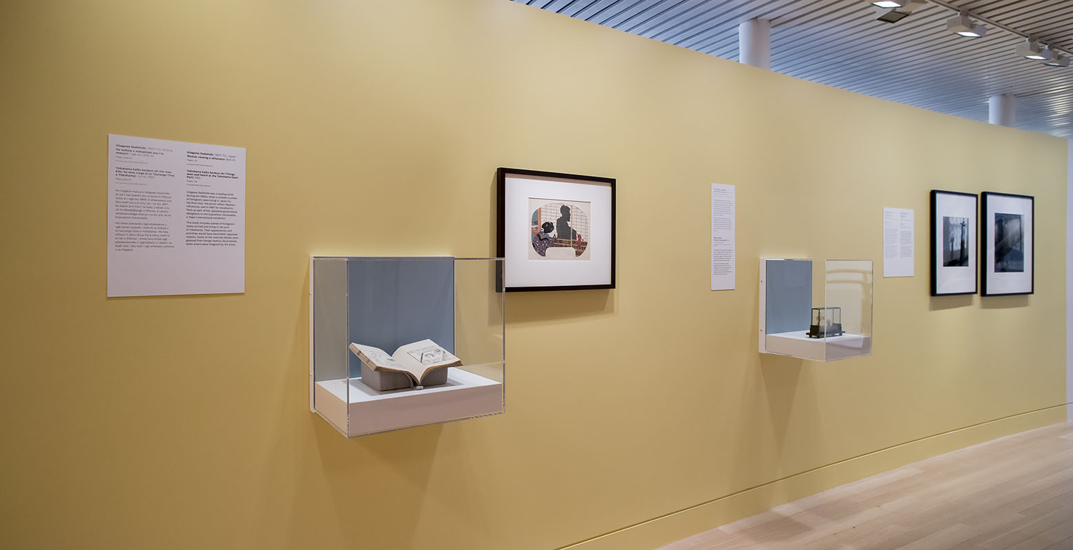 Two photographs are hung on a yellow wall, with a book and a wooden Chinese garden scene housed in display cases