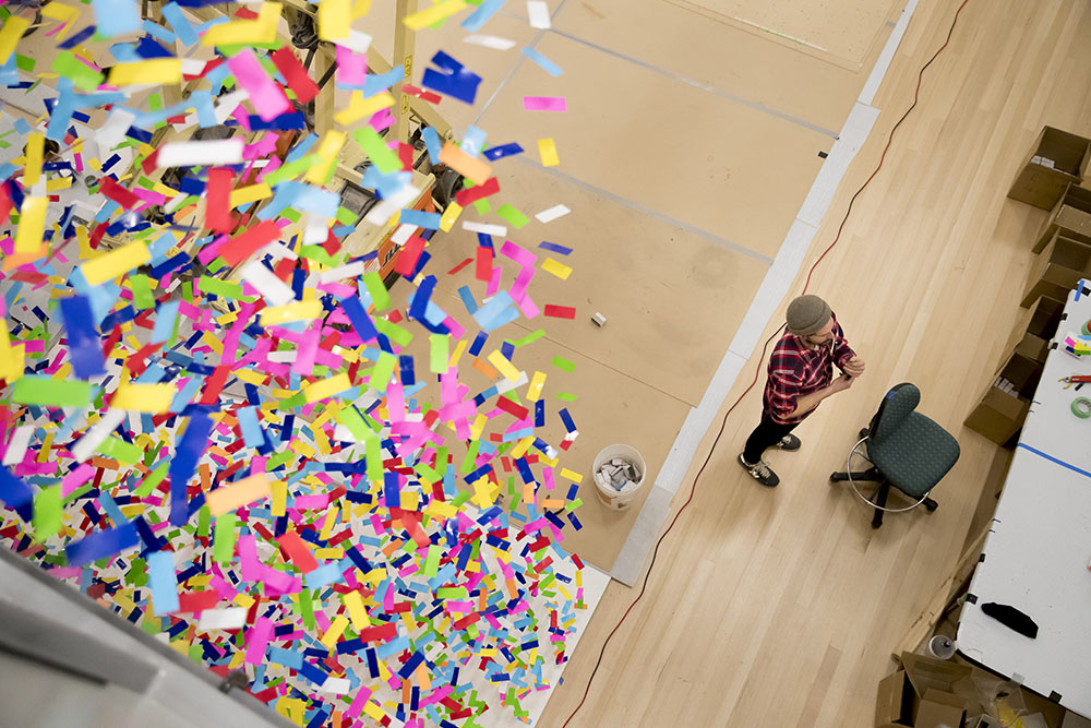 View from above looking down at part of Finale: Bouquet already hung. To the right, in a space yet to be installed stands a man in front of a chair and lots of carton containing the boxes of strands for the work