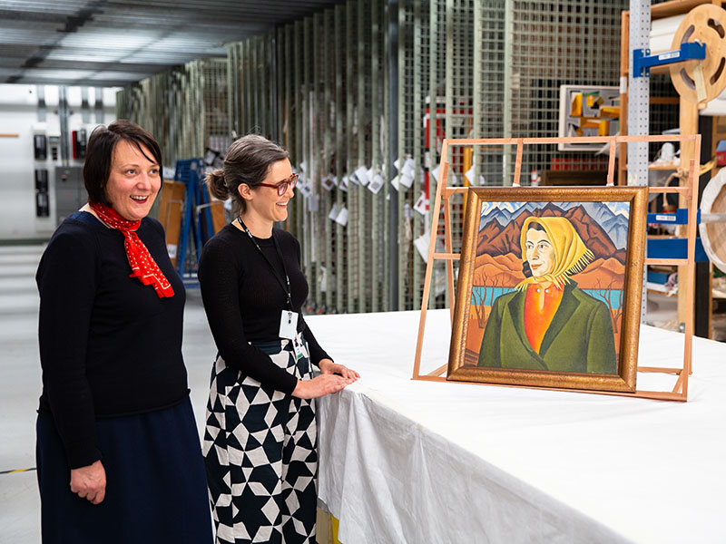 Two women in a painting store room look at a painting on display on a table