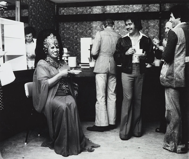 Carmen sits with a bowl while several men stand around with coffee cups and bowls in front of what appears to be self-serve breakfast