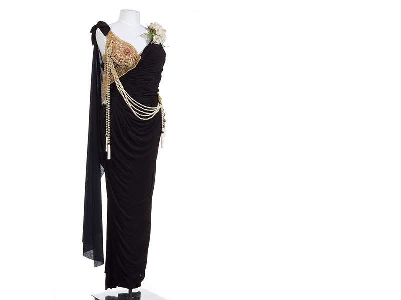 Black sleeveless dress with cape and pearls hanging off the shoulder strap and a corsage and multiple decorative pieces