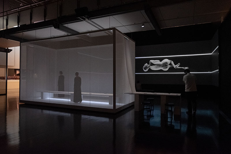 A man walks around a space that contains a box with two women inside and a massive projection on the wall of a woman contorting her body