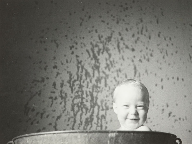 A baby grins, he's poking his head above a bathtub. There's water splashes on the walls.