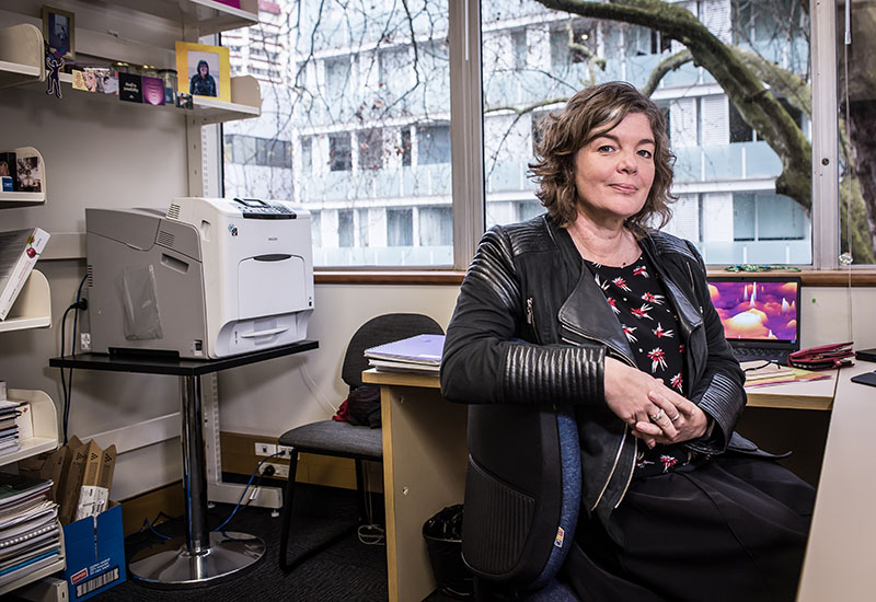 Juliet sits at her desk, with her arm over the back of the chair, in her office. Behind her is a large printer and shelves with books and framed photos