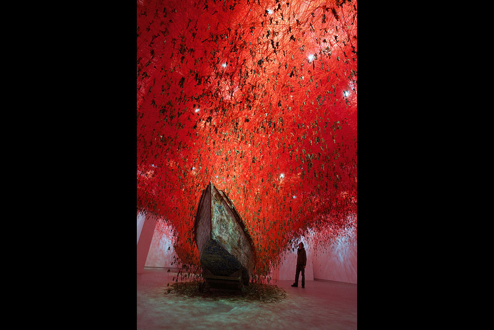 Large network of red thread fills a room from an old boat on the ground to the ceiling. In the threads hang old keys