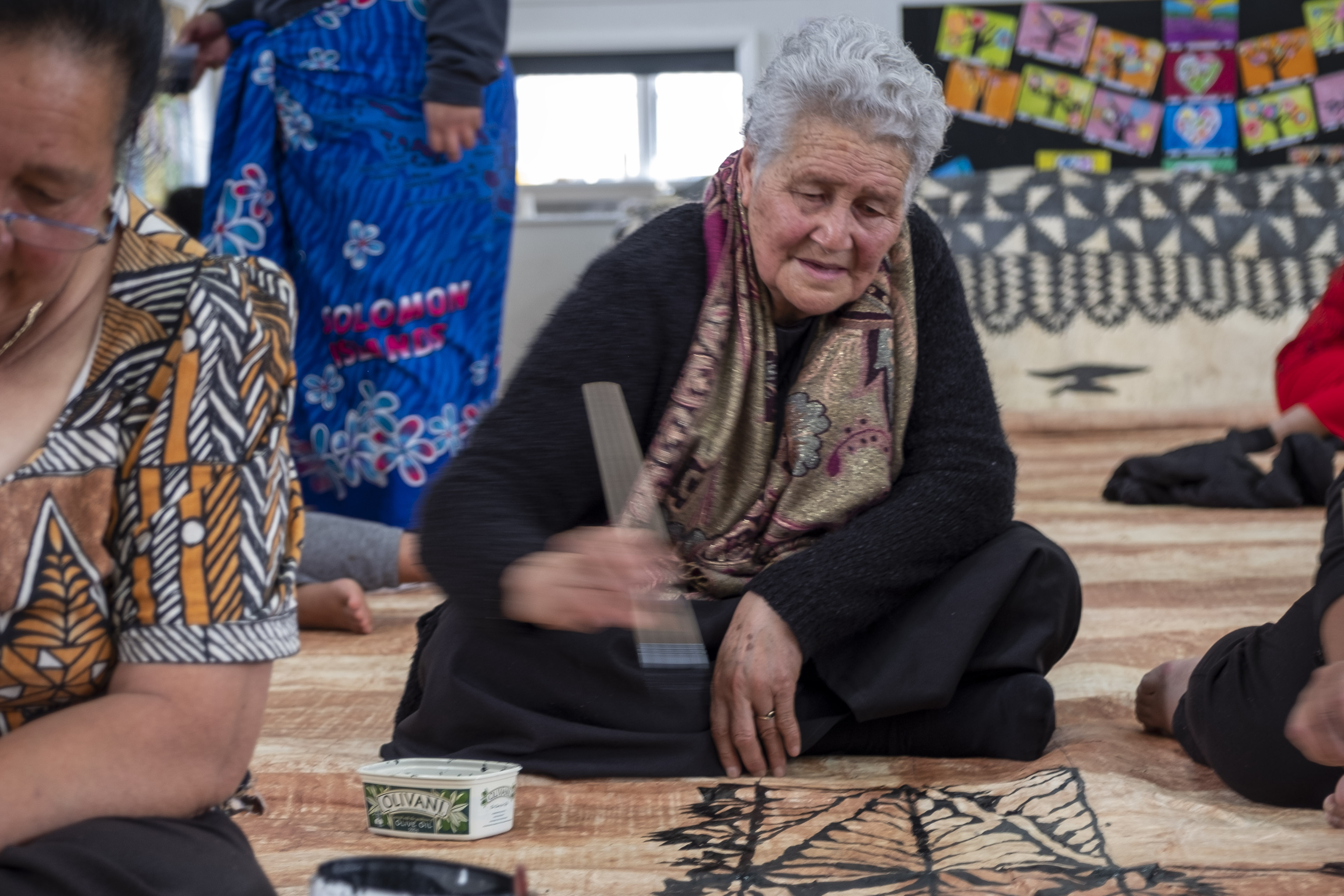 A woman painting black ink on a tapa cloth on the floor.