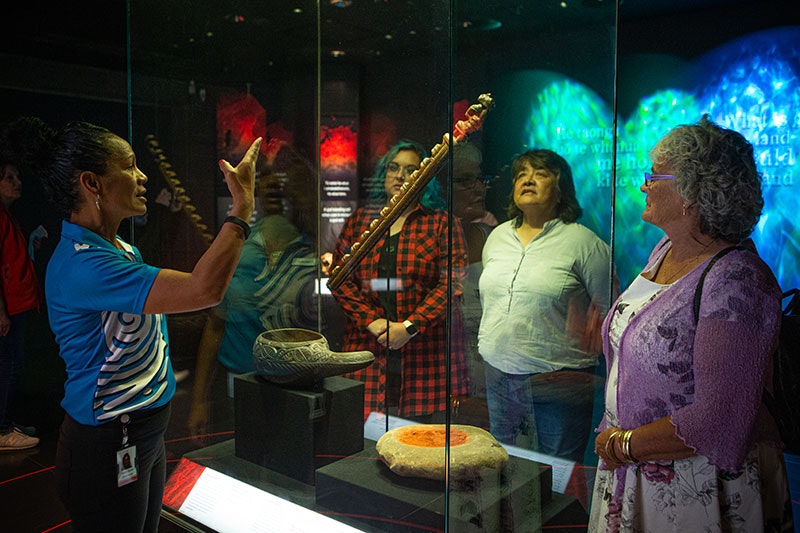 A Te Papa host points at an object in a horizontal case while visitors look on