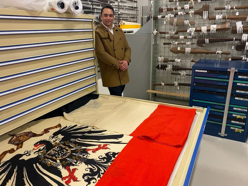 A man in a khaki coat stands against a set of storage drawers. On of the drawers is open and on display in it is an Imperial German flag featuring stripes of equal size in black, white, and red. In the middle is a picture of a large black eagle