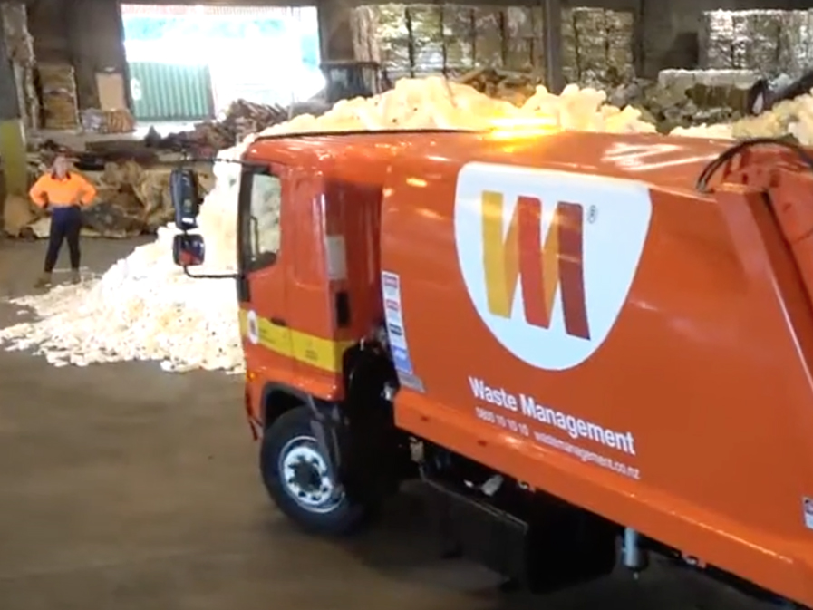 A waste management truck is in a shed with mounds of empty plastic milk bottles and a person standing in the background