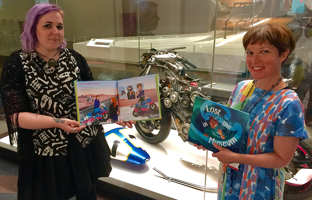 Two women in front of a museum case. Both are holding books