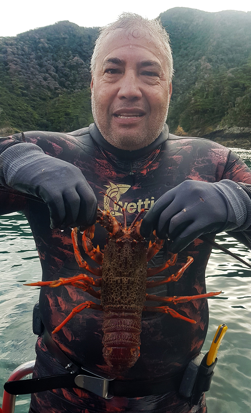 A man in a wetsuit is holding a crayfish up in front of himself. He is looking at the camera.