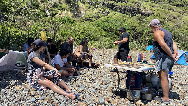 Two men standing and several people sitting on a rocky beach. One of the men standing is talking to the people sitting down.