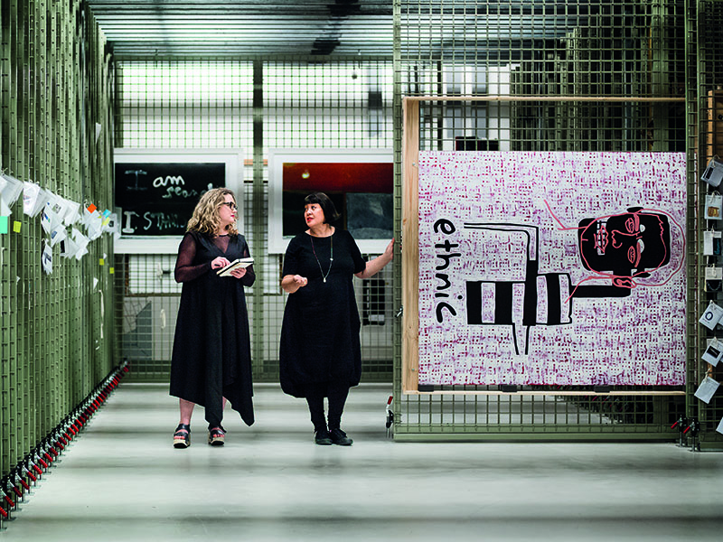 Sarah Farrar and Megan Tamati-Quennell in the paintings storeroom, with a painting on a rail pulled out and on display, with two other paintings in the background