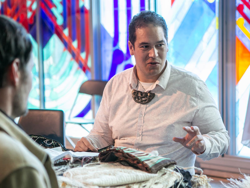 Isaac sitting at a table with kākahu (Māori cloaks) in front of him