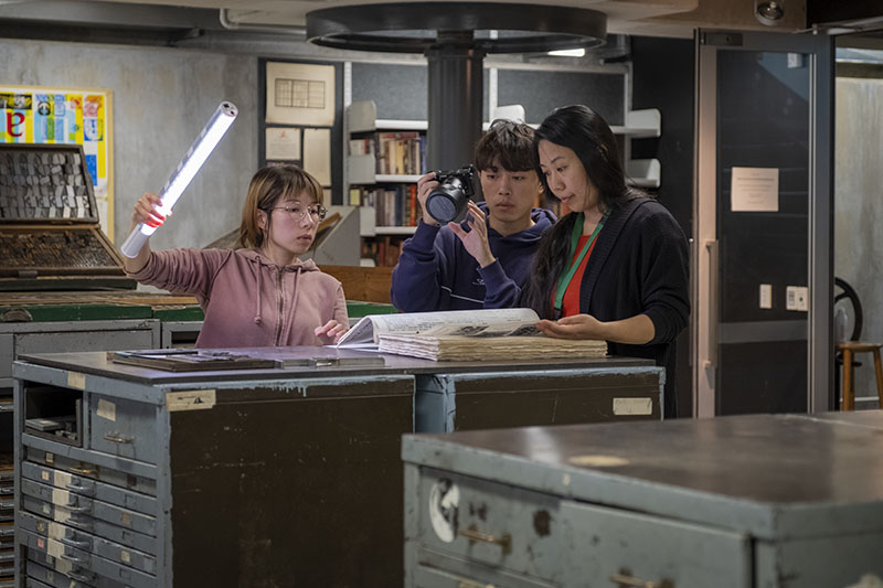 Three people look at a large book on top of a shelving unit. One person holds a light to illuminate the book, one person is filming the scene, and another person is looking through the book