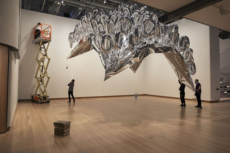 Three people on the floor and one person in a scissor lift install a large bunch of silver helium balloons in a large room