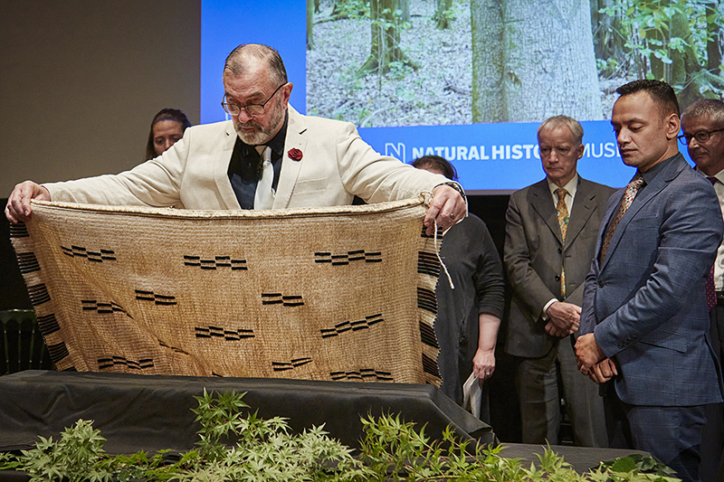 A man holds a cloak out over a cloth-covered box. There are several people standing in the background looking on.