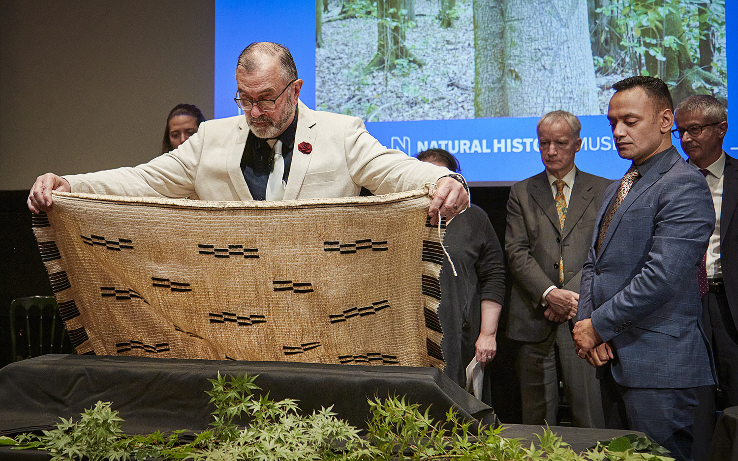 A man lays a woven cloak over a material-covered box while several people behind him look on