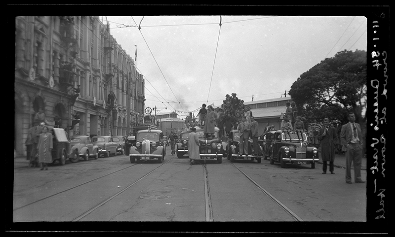 A crowd gathering with some people standing on cars to get a good view of a cavalcade (out of shot)