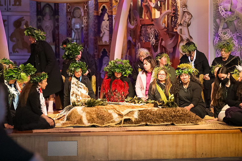 A group of woman sit on a stage surrounding ancestral remains, which are covered by feathered cloaks