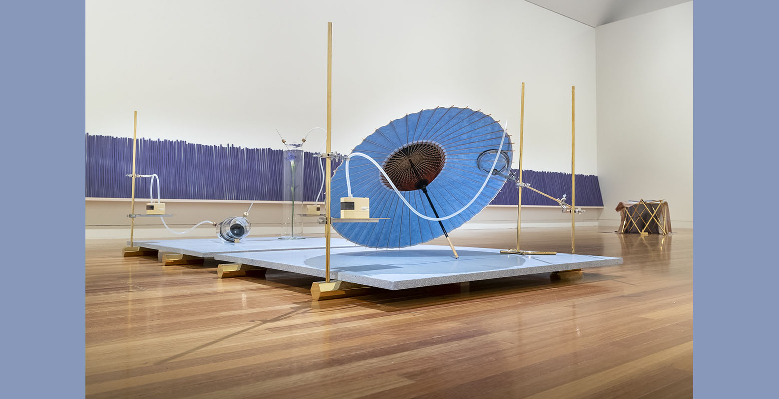 A platform sits on gold-coloured hexagonal feet. On the platform rests an iris-coloured Japanese umbrella, a camera lens and an iris flower in seperate glass jars. In the background a row of large purple incense sticks against the wall.