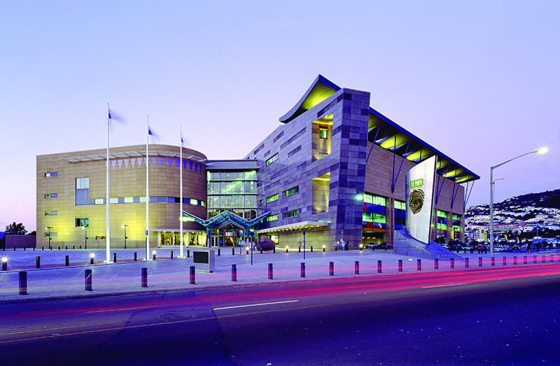 A large building that has oddly shaped walls photographed at dusk. There are three flags outside the entrance.