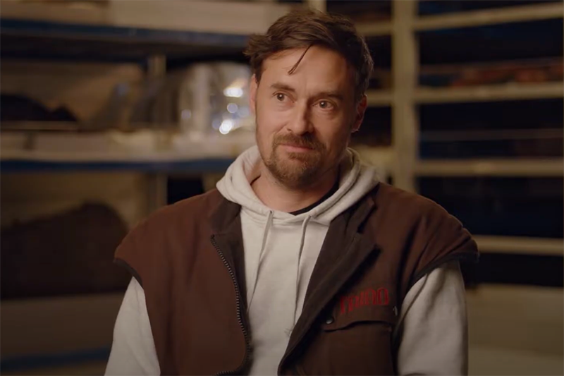 A man with a beard is looking off camera at someone interviewing him. He is in a museum storeroom.