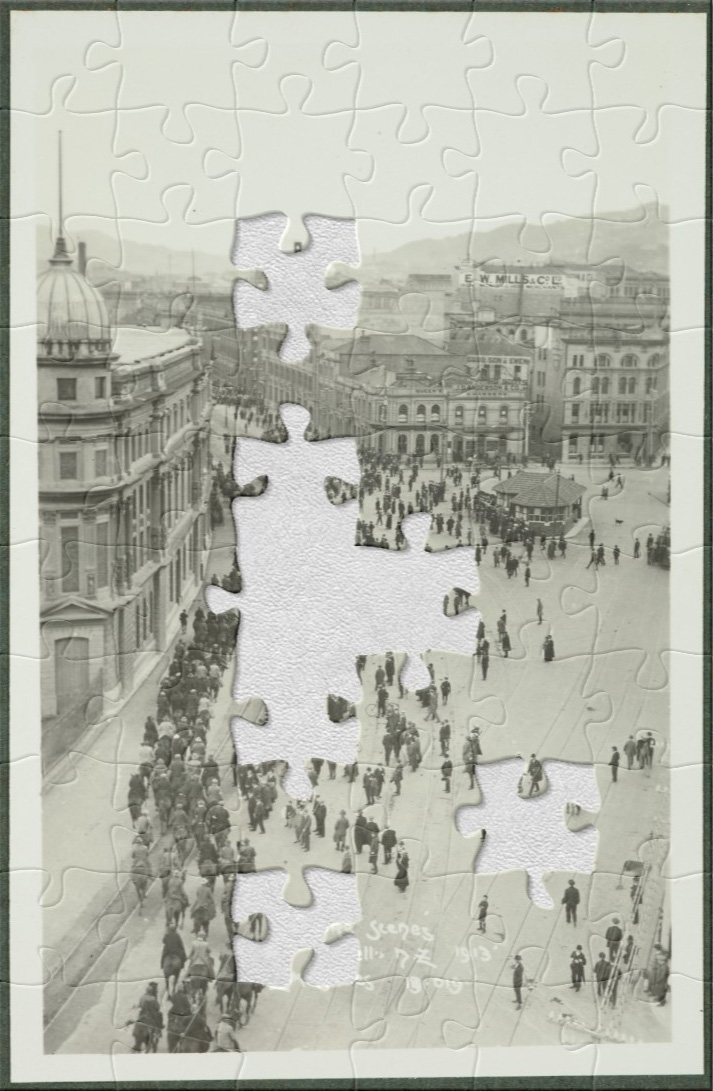 Black and white photo of people on horses and people standing in a row on a wide street with buildings in the background