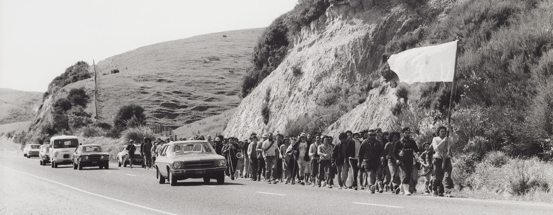 Black and white photo of a large group of people marching alongside a rural road. The person at the front is carrying a large flag