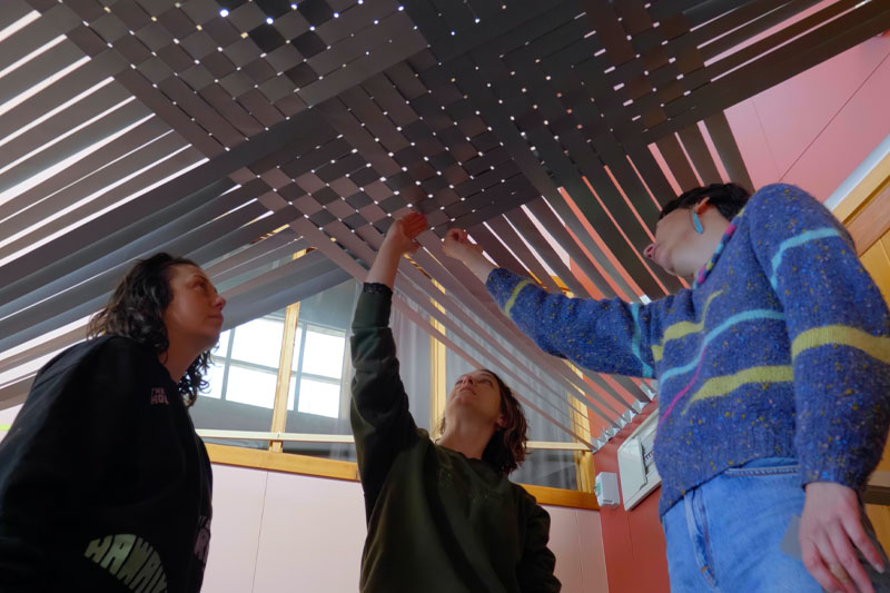 Still from a video. Shows three women underneath a large-scale weaving that they are working on, looking at its finer details