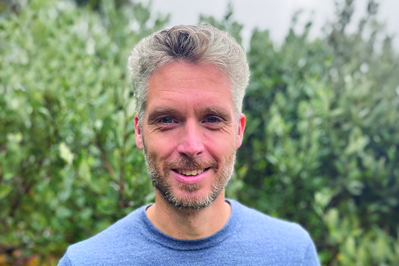 Head and shoulders of a white man with grey hair and a blue jumper. He is standing in front of green shrubbery.