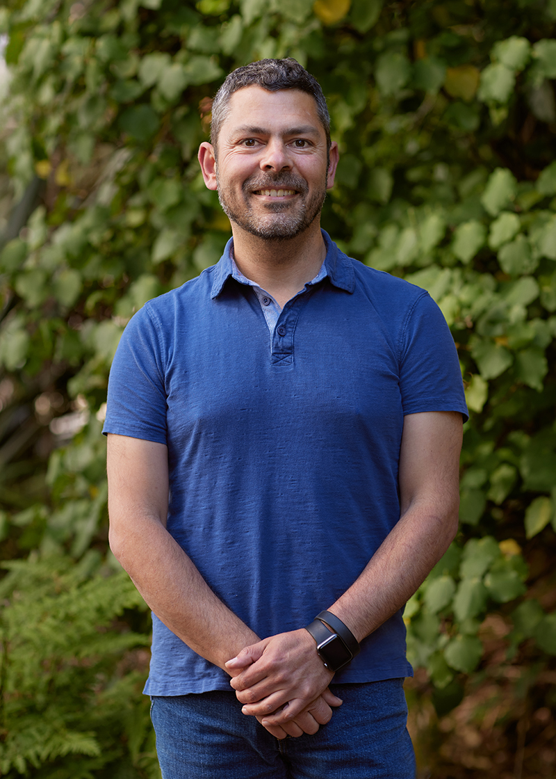 A man with dark hair and a bright blue t-shirt is smiling at the camera