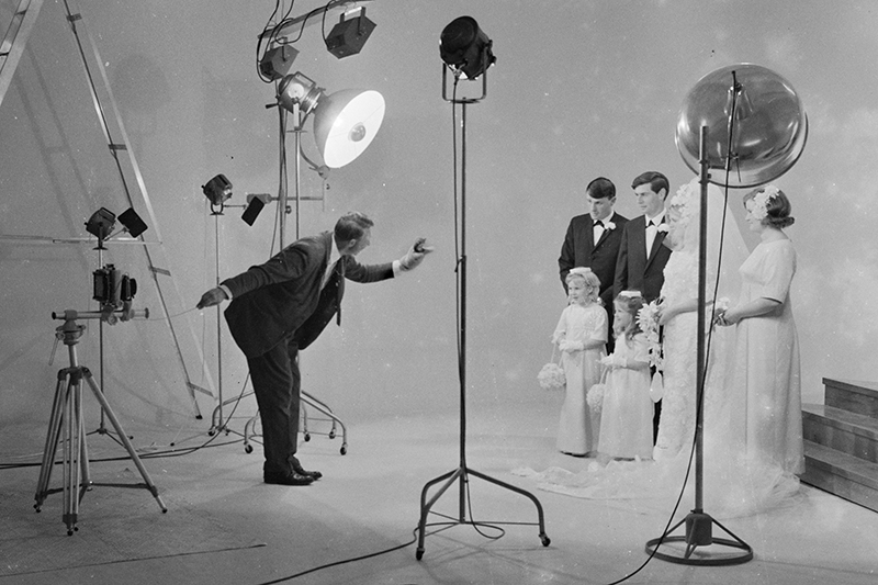 A photographer has a puppet on one hand to entertain children while his other hand is operating a camera to take photos of a wedding party. The room is surrounded in lights and props.