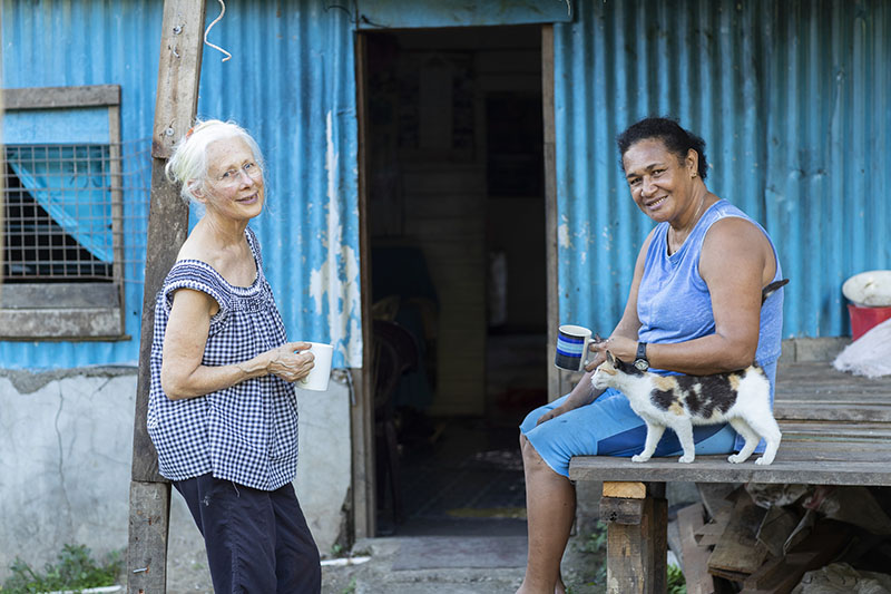 Two women standing in front of a blue corrugated iron shed. They are both holding cups of tea and looking at the camera. There is a cat beside one of them.