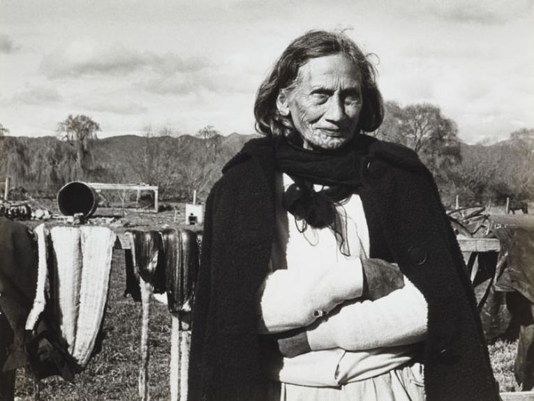 A lady with a chin moko stands outside with her arms folded