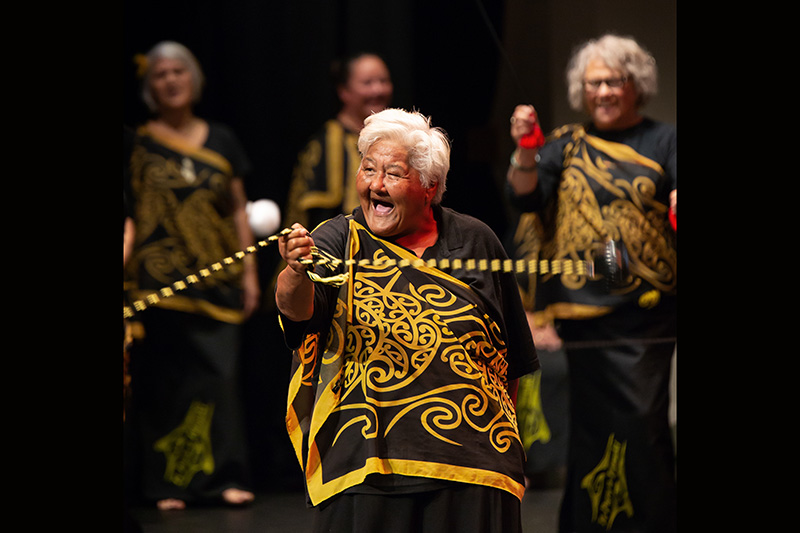 An older woman in a yellow and black top is spinning long poi on stage with other older people.